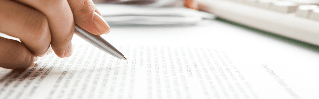 A person's hand holding a pen over a printed document, with focus on the text and a blurred background featuring a keyboard and papers