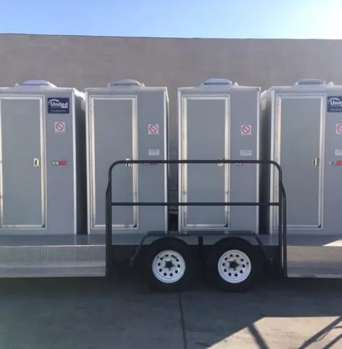 A row of portable toilets neatly arranged alongside a black railing and a small trailer with two wheels, parked against a concrete wall under clear skies