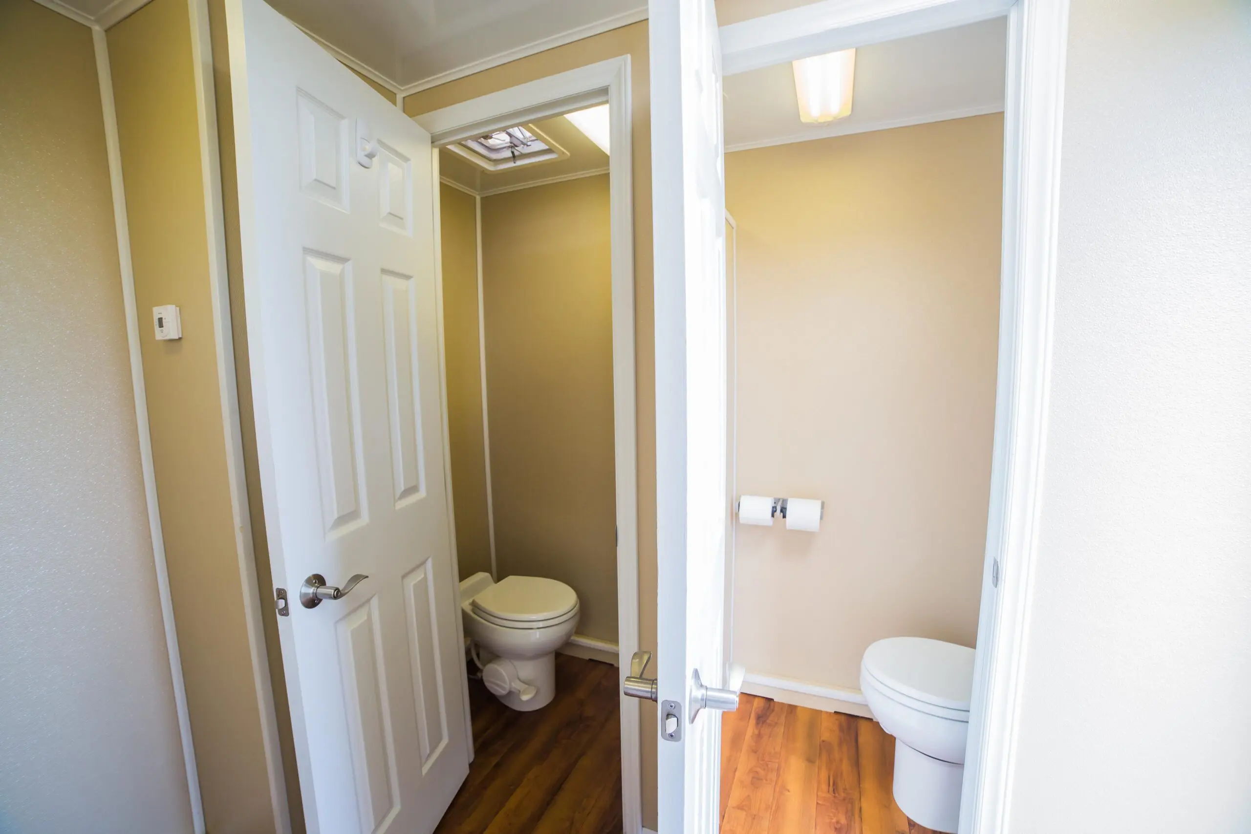 Interior view of a restroom trailer seen through an open door, featuring two bathroom stalls with white toilets and white wooden doors