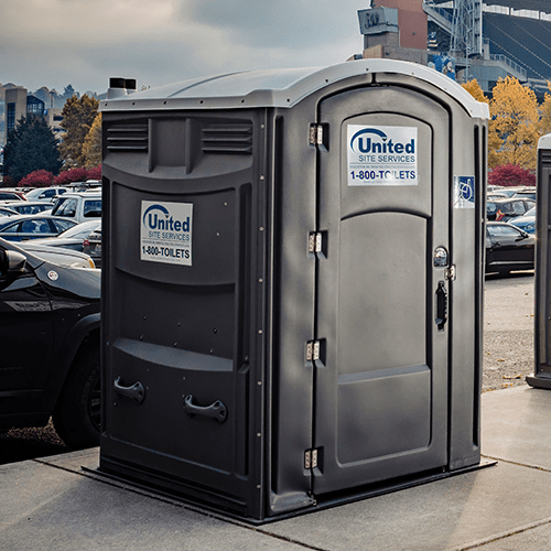 A portable ADA restroom in a stadium parking lot, with cars and stadium seating in the background