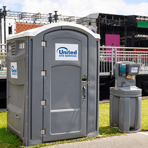 A portable restroom with a hand sanitizer dispenser beside it, stationed near a concert stage set-up in a grassy area under a cloudy sky