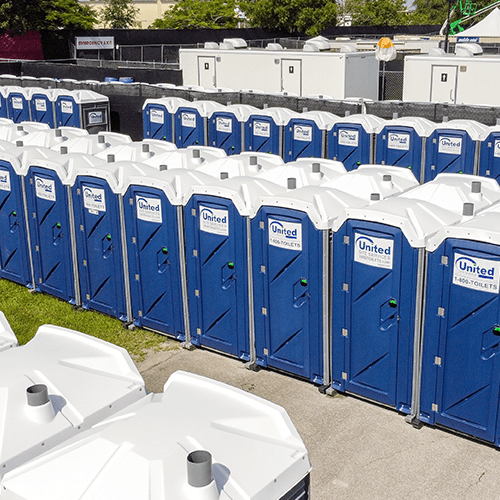 Row of blue portable toilets neatly arranged in an outdoor area, with some white roofs of other units visible in the foreground