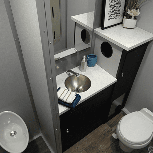 A portable restroom trailer featuring a stainless steel sink, gray countertops, black cabinets, and a white toilet