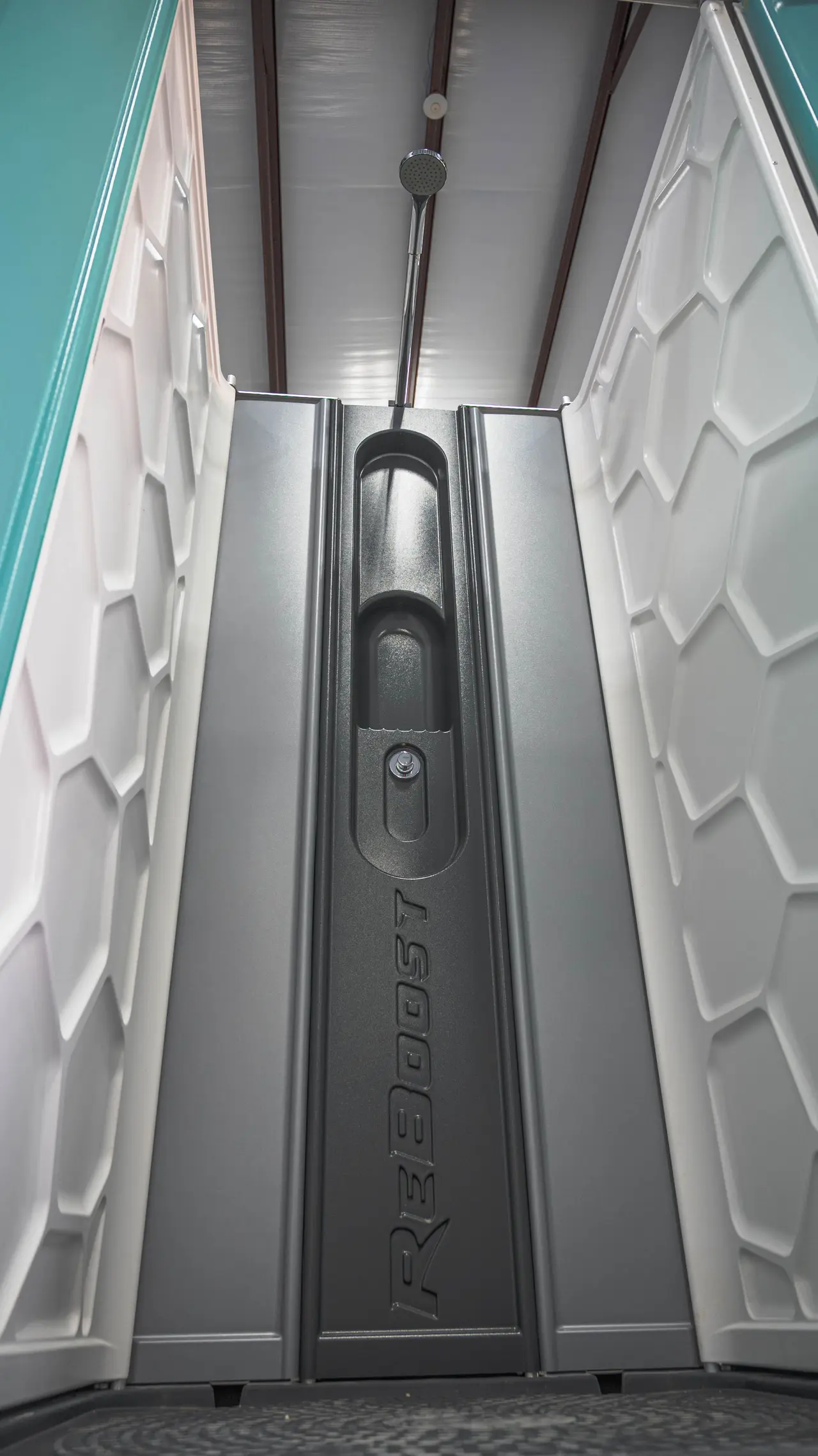 An interior view of a mobile shower unit, focusing on the textured gray door and latch with a hexagonal pattern on the walls, under artificial lighting