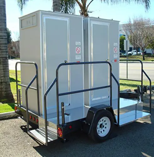 Portable restroom trailer with two doors, mounted on a trailer with a single axle and a small staircase, parked on a street by a grassy area