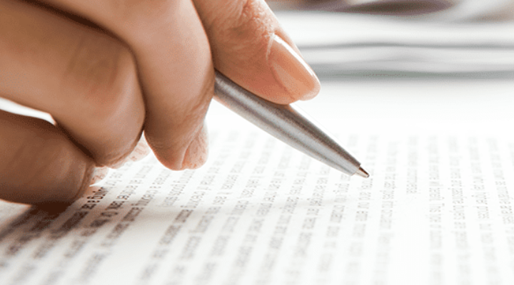 A person's hand holding a pen over a printed document, with focus on the text and a blurred background featuring a keyboard and papers