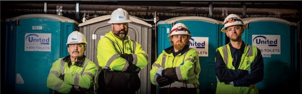 Four United Site Services workers confidently standing in front of portable restrooms, wearing hard hats and High Visibility Vests, with arms crossed