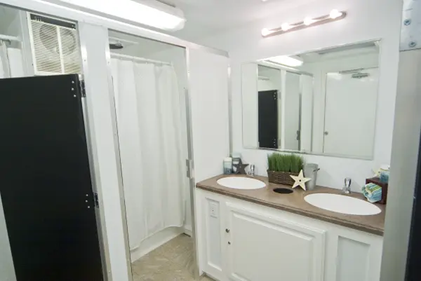 A shower trailer interior with a large mirror above a double sink vanity, white cabinets, a dark shower curtain, and decorative items under bright lights
